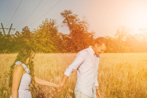 若いカップルの愛の手を握り、干し草の畑で笑顔。男は女を前進させる。夏にポーズを取る光の服で美しいロマンチックなカップルの屋外の肖像画。レンズフレア効果 - 写真・画像