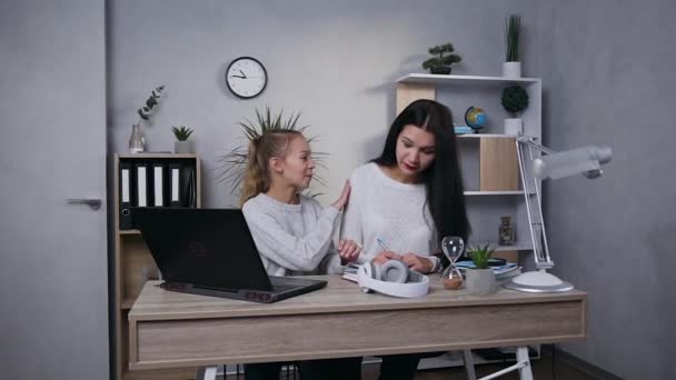 Attractive brunette mother writing in the notebook while her smiling teen daughter working with computer and asking mother to help - Кадри, відео