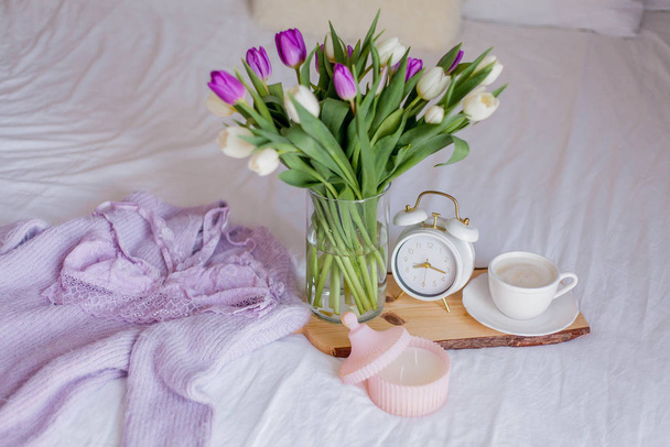 Een boeket tulpen in een glazen vaas, een witte wekker, een kaars, een briefje, een cappuccino mok op een houten plank staan op het bed. Damesondergoed van kant. Ontbijt op bed. - Foto, afbeelding