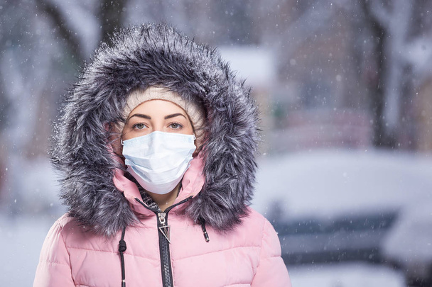 Portret kobiety w różowym kolorze chodzącej zimą ulicą w śniegu w masce ochronnej chroniącej przed chorobami zakaźnymi. Ochrona przed przeziębieniem, grypą, zanieczyszczeniem powietrza. Koncepcja zdrowia - Zdjęcie, obraz