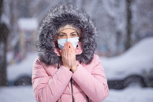 Portrait of woman in pink walking down the street in winter under snowfall in a protective mask to protect against infectious diseases. Protection against colds, flu, air pollution. Health concept - 写真・画像