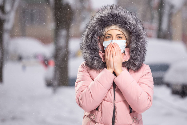 Portrait de femme en rose marchant dans la rue en hiver sous la neige sous un masque protecteur pour se protéger contre les maladies infectieuses. Protection contre le rhume, la grippe, la pollution atmosphérique. Concept de santé
 - Photo, image