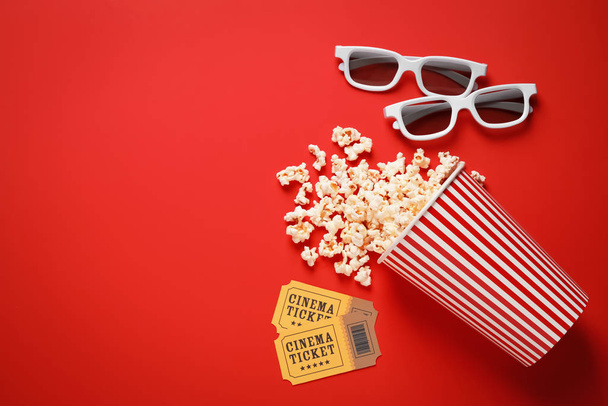 Overturned paper cup with delicious popcorn, cinema tickets and 3D glasses on red background, flat lay. Space for text - 写真・画像