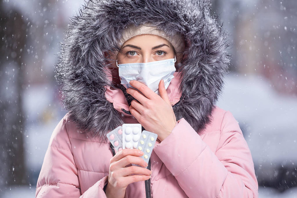 Porträt eines schönen Mädchens in rosa Jacke und medizinischer Schutzmaske, das weiße, rosa und gelbe Pillen gegen Erkältungen und Grippe in den Händen hält. Winterliches Straßenporträt einer Frau unter Schneefall - Foto, Bild