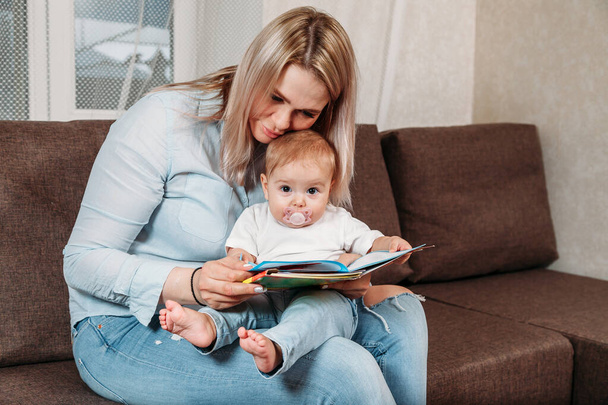 happy mother read a book to child girl indoors sitting on the couch - Фото, изображение