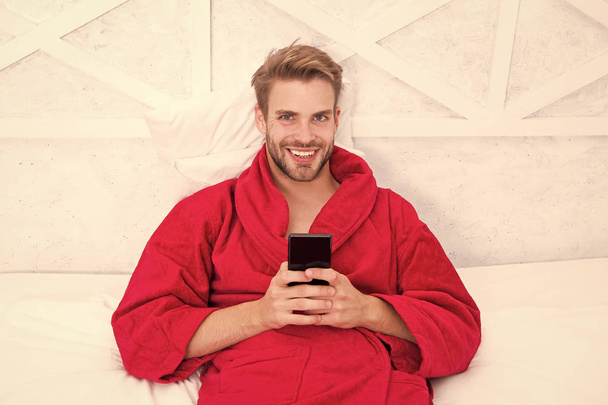 Taşınabilir bir yaşam tarzı. Yatakta mobil cihaz kullanan neşeli bir adam. Elinde cep telefonuyla gülümseyen yakışıklı adam. Mobil iletişim ve evdeki kablosuz teknolojiler - Fotoğraf, Görsel