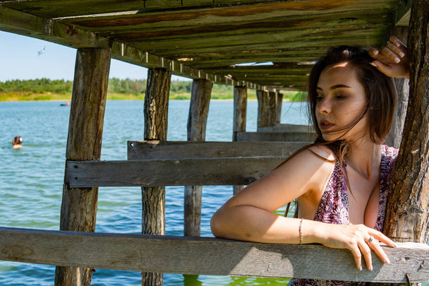 Mujer morena posando en el muelle de madera del lago. Mujer en traje de verano con la espalda vuelta a la pasarela de madera del lago Neusiedl. Reflejos solares en la casa de botes en el fondo
. - Foto, Imagen