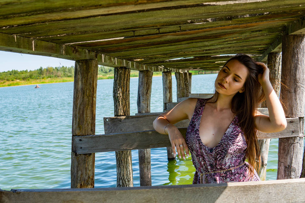 Mujer morena posando en el muelle de madera del lago. Mujer en traje de verano con la espalda vuelta a la pasarela de madera del lago Neusiedl. Reflejos solares en la casa de botes en el fondo
. - Foto, imagen