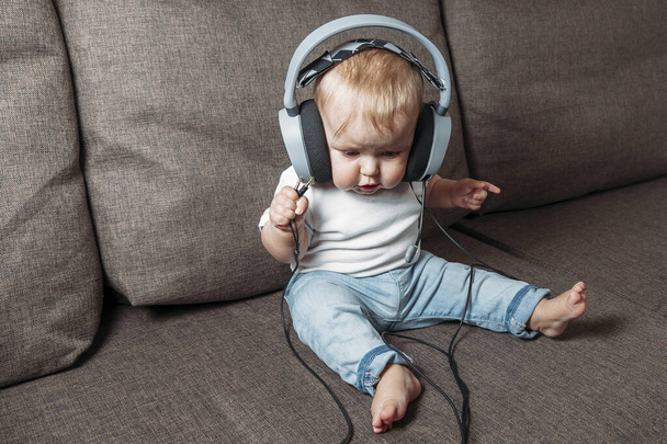 little child listening music with headphones on his head sitting on sofa at home  - Foto, afbeelding