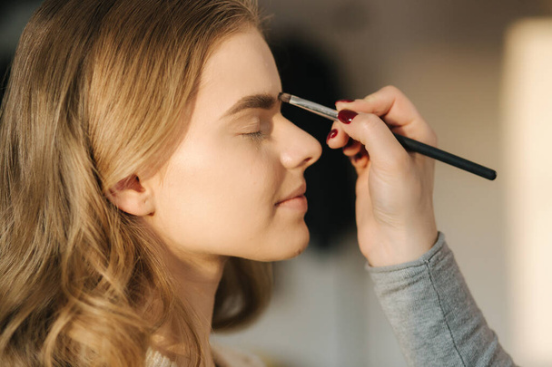 Makeup artist work in her beauty studio. Portrait of Woman applying by professional make up master. Beautiful make up artist start making a makeup for blond hair model - Photo, Image
