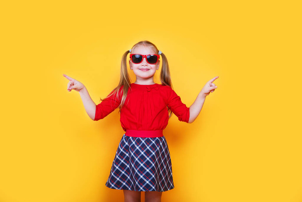 Little girl wearing sunglasses smiling and pointing to the side with thumb up. - Photo, image