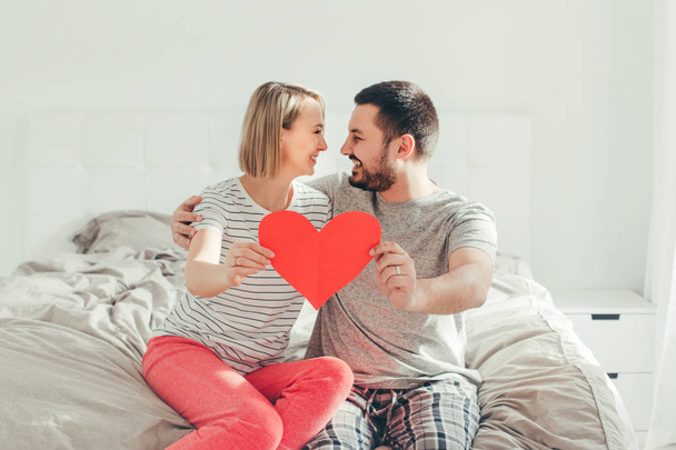 Happy strong marriage. Happy funny couple in love holding red paper heart. Heterosexual family man and woman sitting on bed in bedroom at home hugging. Real people authentic lifestyle.  - 写真・画像