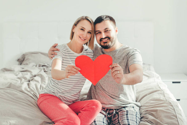 Happy strong marriage. Happy funny couple in love holding red paper heart. Heterosexual family man and woman sitting on bed in bedroom at home hugging. Real people authentic lifestyle.  - Foto, immagini