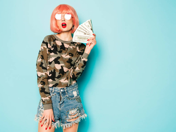 Young beautiful hipster bad girl in trendy red summer clothes and earring in her nose.Sexy carefree smiling woman sitting in studio in pink wig near blue wall.Positive model holding dollars in hands - Φωτογραφία, εικόνα