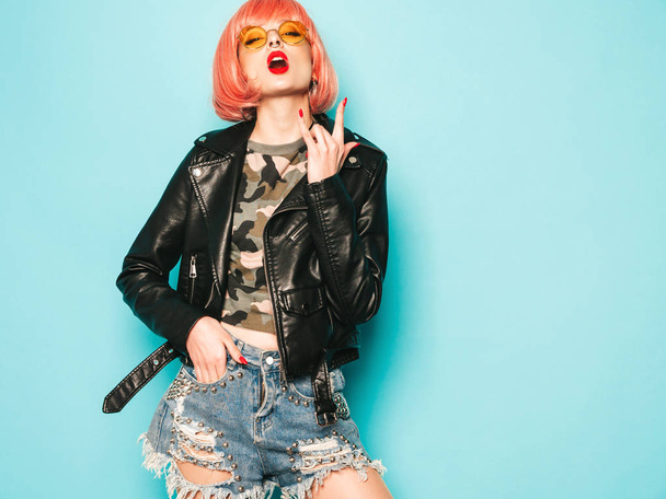 Young beautiful hipster bad girl in black leather jacket and earring in nose.Sexy carefree  woman posing in studio in pink wig near blue wall.Confident model in sunglasses.Shows rock and roll sign - Fotó, kép