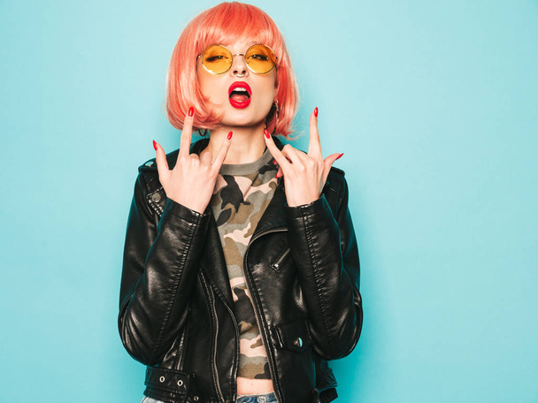 Young beautiful hipster bad girl in black leather jacket and earring in nose.Sexy carefree  woman posing in studio in pink wig near blue wall.Confident model in sunglasses.Shows rock and roll sign - Φωτογραφία, εικόνα