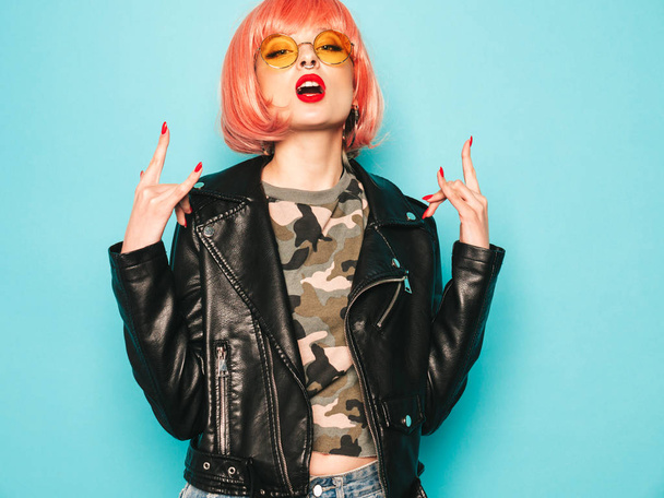 Young beautiful hipster bad girl in black leather jacket and earring in nose.Sexy carefree  woman posing in studio in pink wig near blue wall.Confident model in sunglasses.Shows rock and roll sign - Foto, Bild