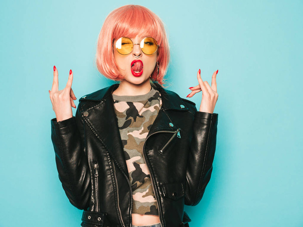 Young beautiful hipster bad girl in black leather jacket and earring in nose.Sexy carefree  woman posing in studio in pink wig near blue wall.Confident model in sunglasses.Shows rock and roll sign - Fotó, kép
