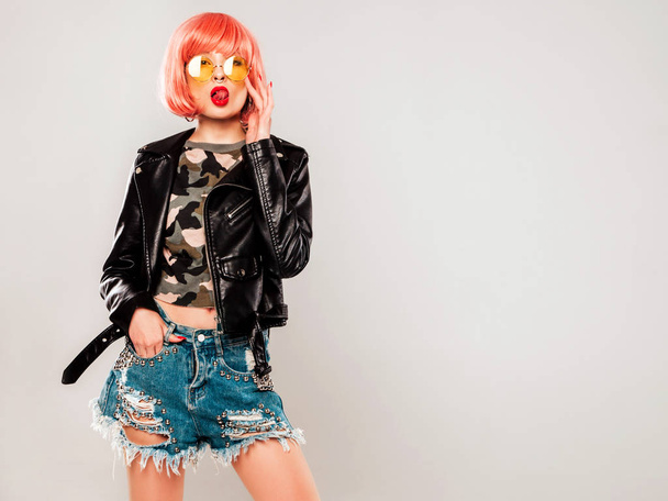 Portrait of young beautiful hipster bad girl in black leather jacket and earring in her nose.Sexy carefree smiling woman sitting in studio in pink wig near gray wall.Confident model in sunglasses - 写真・画像