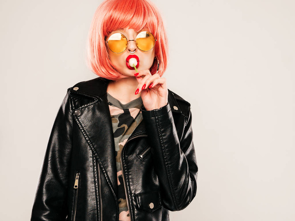 Closeup lips of young beautiful hipster bad girl in trendy black leather jacket and earring in her nose.Sexy carefree smiling woman posing in studio in pink wig.Positive model licking round candy - Foto, Imagen