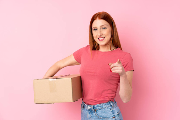 Young redhead woman over isolated pink background holding a box to move it to another site while pointing to the front - Фото, изображение