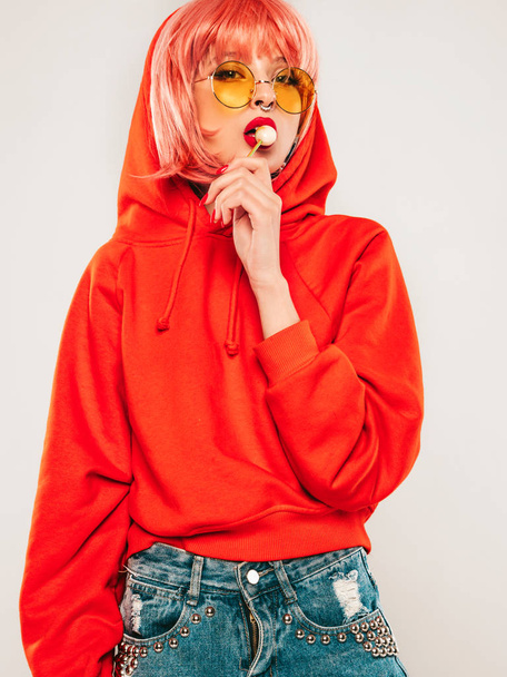 Young beautiful hipster bad girl in trendy red summer red hoodie and earring in her nose.Sexy carefree woman posing in studio on gray background in wig.Hot model licking round sugar candy - Фото, зображення