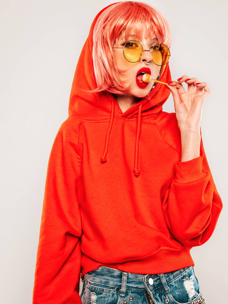 Young beautiful hipster bad girl in trendy red summer red hoodie and earring in her nose.Sexy carefree woman posing in studio on gray background in wig.Hot model licking round sugar candy - 写真・画像