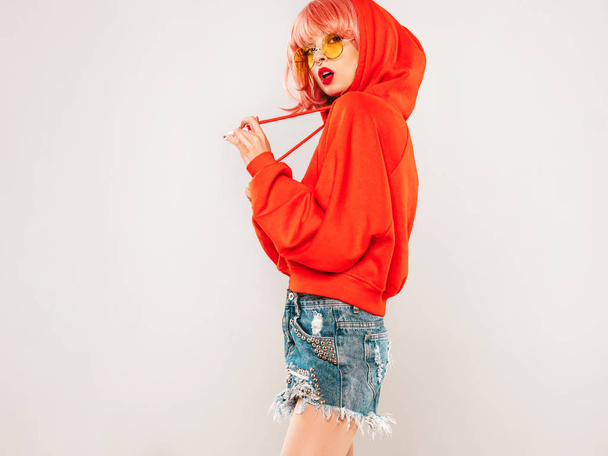 Young beautiful hipster bad girl in trendy red summer hoodie and earring in her nose.Sexy carefree woman in pink wig posing in studio on gray background.Hot model in sunglasses - 写真・画像