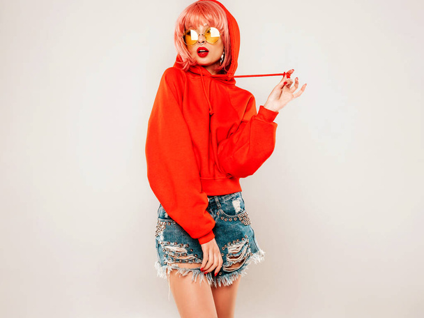 Young beautiful hipster bad girl in trendy red summer hoodie and earring in her nose.Sexy carefree woman in pink wig posing in studio on gray background.Hot model in sunglasses - Фото, изображение