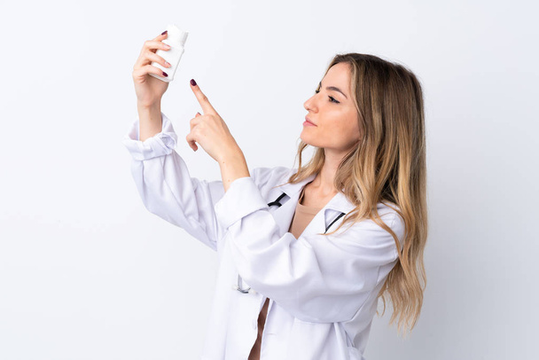Young woman over isolated white background wearing a doctor gown and holding pills - 写真・画像