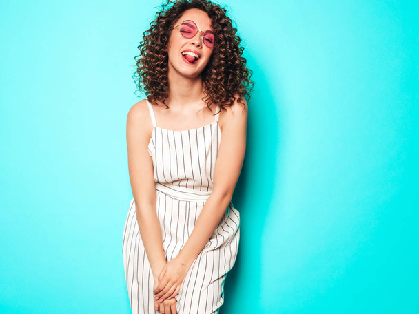 Portrait of beautiful smiling model with afro curls hairstyle dressed in summer hipster clothes.Sexy carefree girl posing near blue wall.Trendy funny and positive woman.Shows tongue - Photo, Image