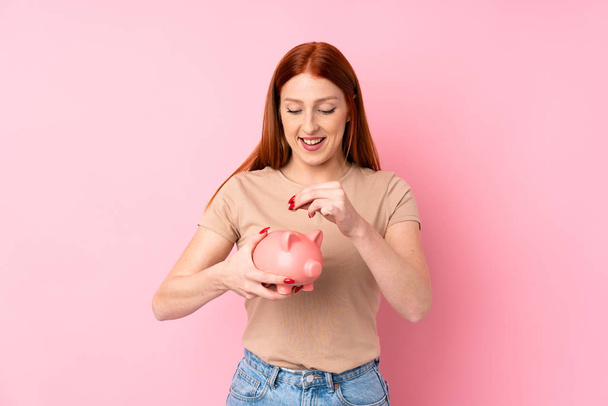 Young redhead woman over isolated pink background holding a big piggybank - Photo, Image