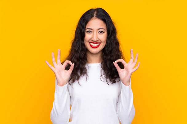 Смешанная женщина расы на изолированном желтом фоне показывает знак ОК с пальцами
 - Фото, изображение