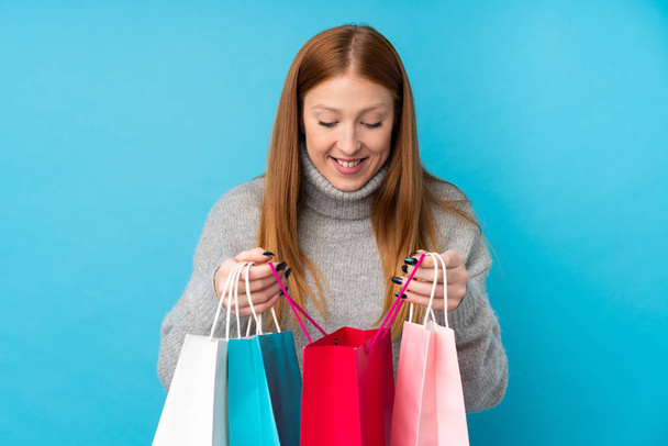 Young redhead woman over isolated blue background holding shopping bags and looking inside it - Foto, imagen