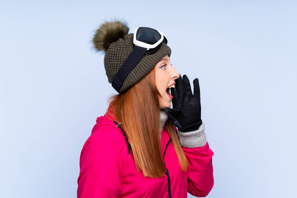 Skier rousse femme avec des lunettes de snowboard sur mur bleu isolé criant avec bouche grande ouverte
 - Photo, image
