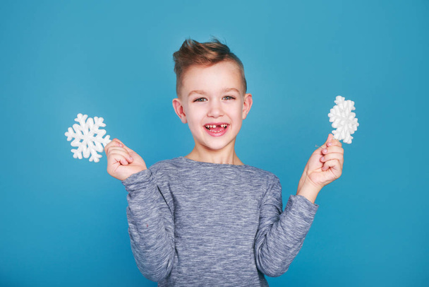 Little smiling boy holding a cardboard snowflake on blue background - Foto, imagen