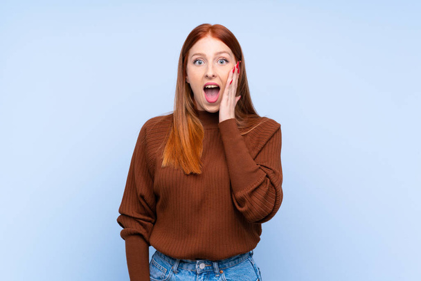 Junge rothaarige Frau vor isoliertem blauen Hintergrund mit Überraschung und schockiertem Gesichtsausdruck - Foto, Bild