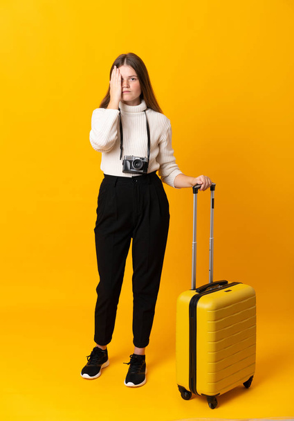 Full body of traveler teenager girl with suitcase over isolated yellow background covering a eye by hand - Fotó, kép