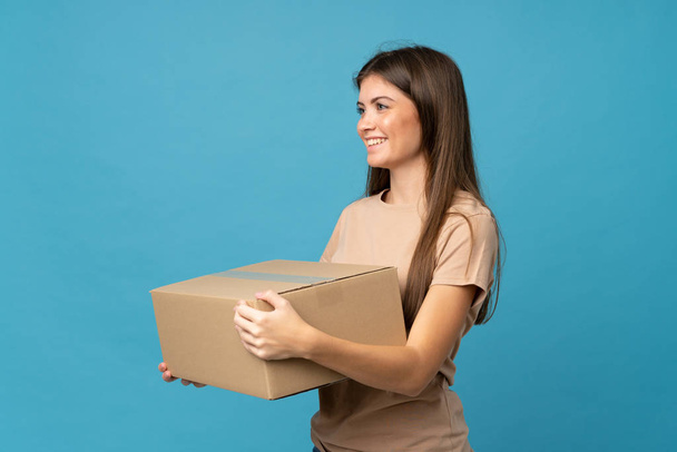 Młoda kobieta na odizolowanym niebieskim tle trzymając pudełko, aby przenieść go do innego miejsca - Zdjęcie, obraz
