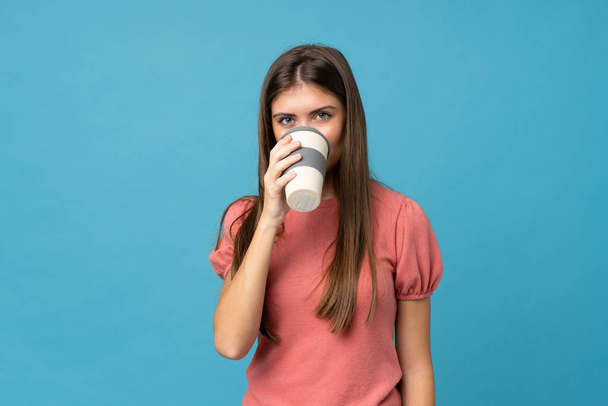 Mujer joven sobre fondo azul aislado sosteniendo y bebiendo un café para llevar
 - Foto, Imagen