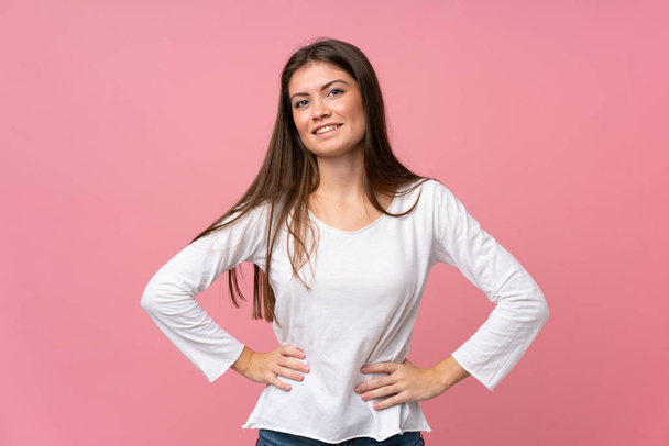 jonge vrouw over geïsoleerde roze achtergrond poseren met armen op heup en glimlachen - Foto, afbeelding
