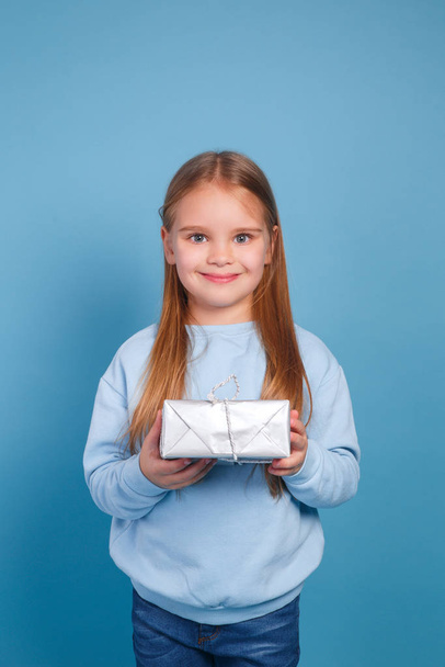 Portrait of a little girl in a warm knitted sweater with gift box on a cyan background - Фото, зображення