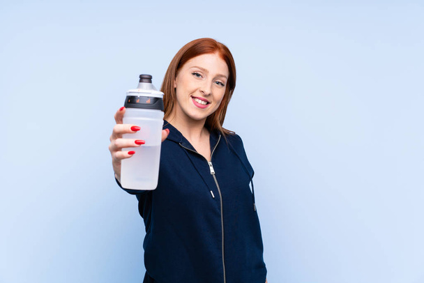 Genç kızıl saçlı sporcu kadın izole edilmiş mavi arka planda spor su şişesiyle - Fotoğraf, Görsel