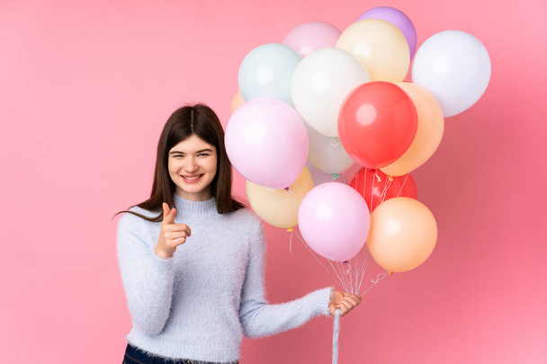 Junge ukrainische Teenager Mädchen, das viele Luftballons über isoliertem rosa Hintergrund hält, zeigt mit dem Finger auf dich - Foto, Bild