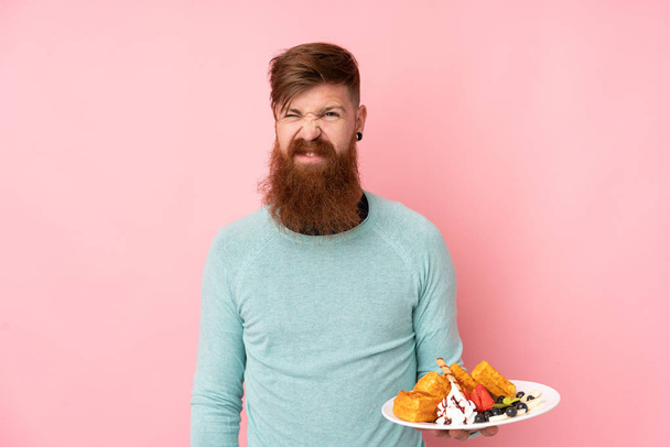 Redhead man with long beard holding waffles over isolated pink background with sad expression - Фото, изображение