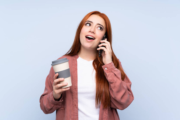 Pelirroja adolescente chica sobre aislado fondo azul celebración de café para llevar y un móvil - Foto, Imagen