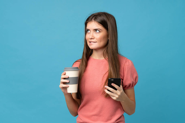 junge Frau über isoliertem blauem Hintergrund mit Kaffee zum Mitnehmen und einem Handy, während sie etwas denkt - Foto, Bild