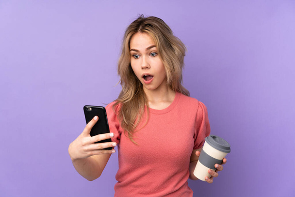 Teenager russische Mädchen isoliert auf lila Hintergrund hält Kaffee zum Mitnehmen und ein Handy - Foto, Bild