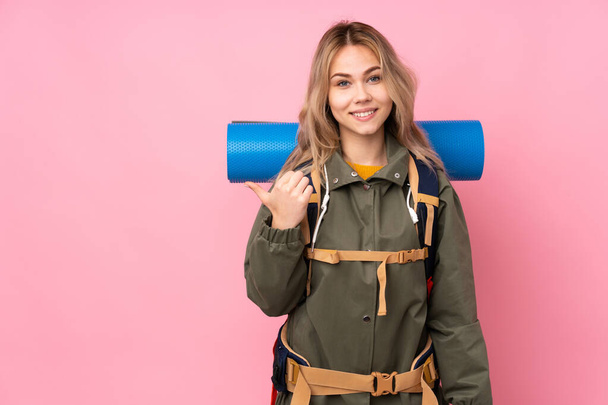 Tiener Russisch bergbeklimmer meisje met een grote rugzak geïsoleerd op roze achtergrond wijzend naar de zijkant om een product te presenteren - Foto, afbeelding