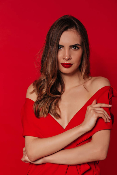 selbstbewusste hübsche Frau mit welligem Haar, die in einem roten Kleid mit roten Lippen vor rotem Hintergrund in die Kamera blickt. Stylisches Frauenporträt  - Foto, Bild